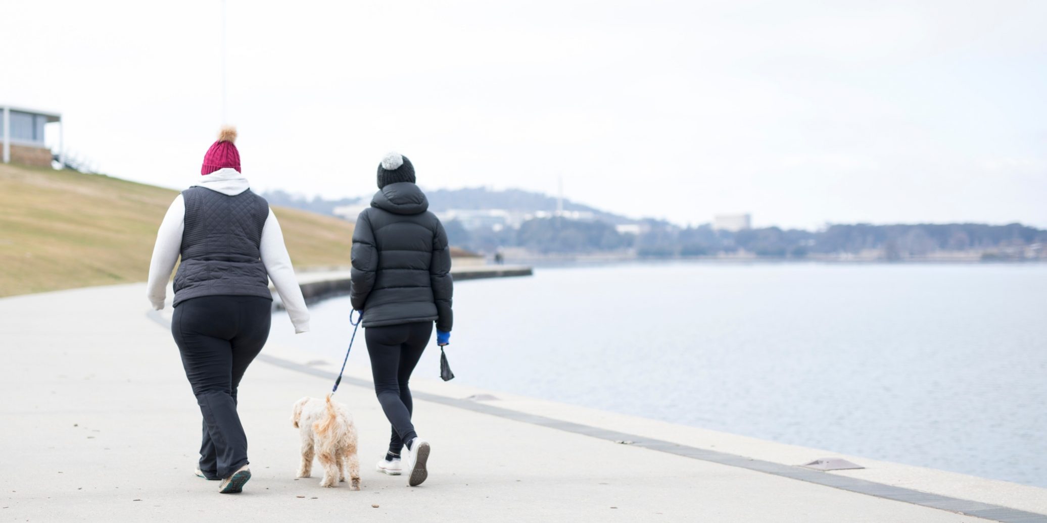 two-friends-rugged-up-in-winter-puffy-jackets-beanies-and-mittens-taking-the-dog-for-a-walk-around_t20_oopKN8